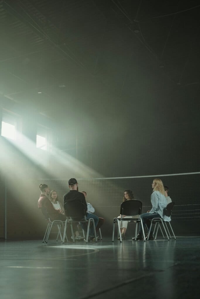 A group of adults in a therapy session, sitting in a dimly lit room with sun rays streaming in.